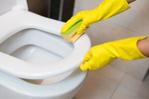 Mani con guanti gialli che tengono sollevata la tavoletta del WC e la puliscono con una spugna